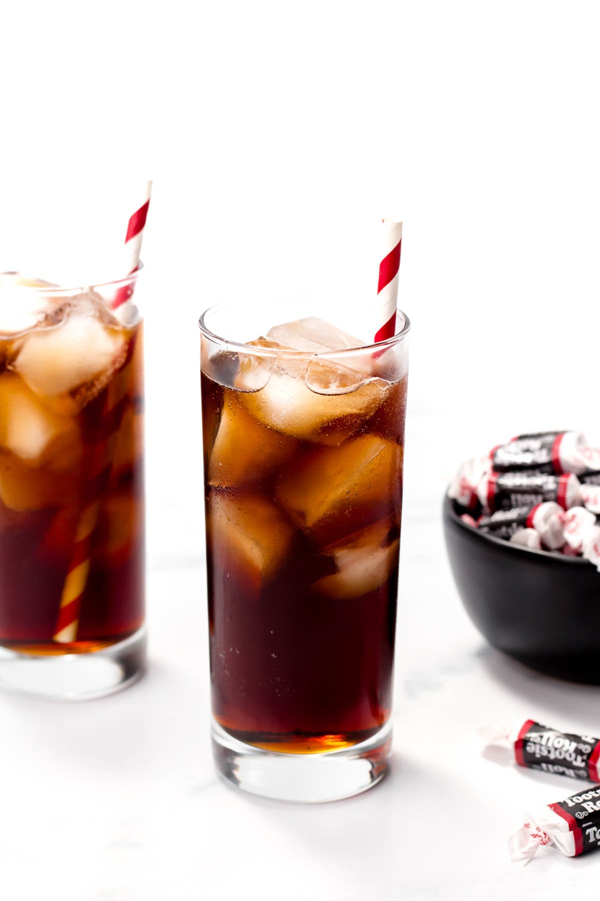 Two glasses of iced cola with striped straws sit on a table next to a bowl of wrapped candies.