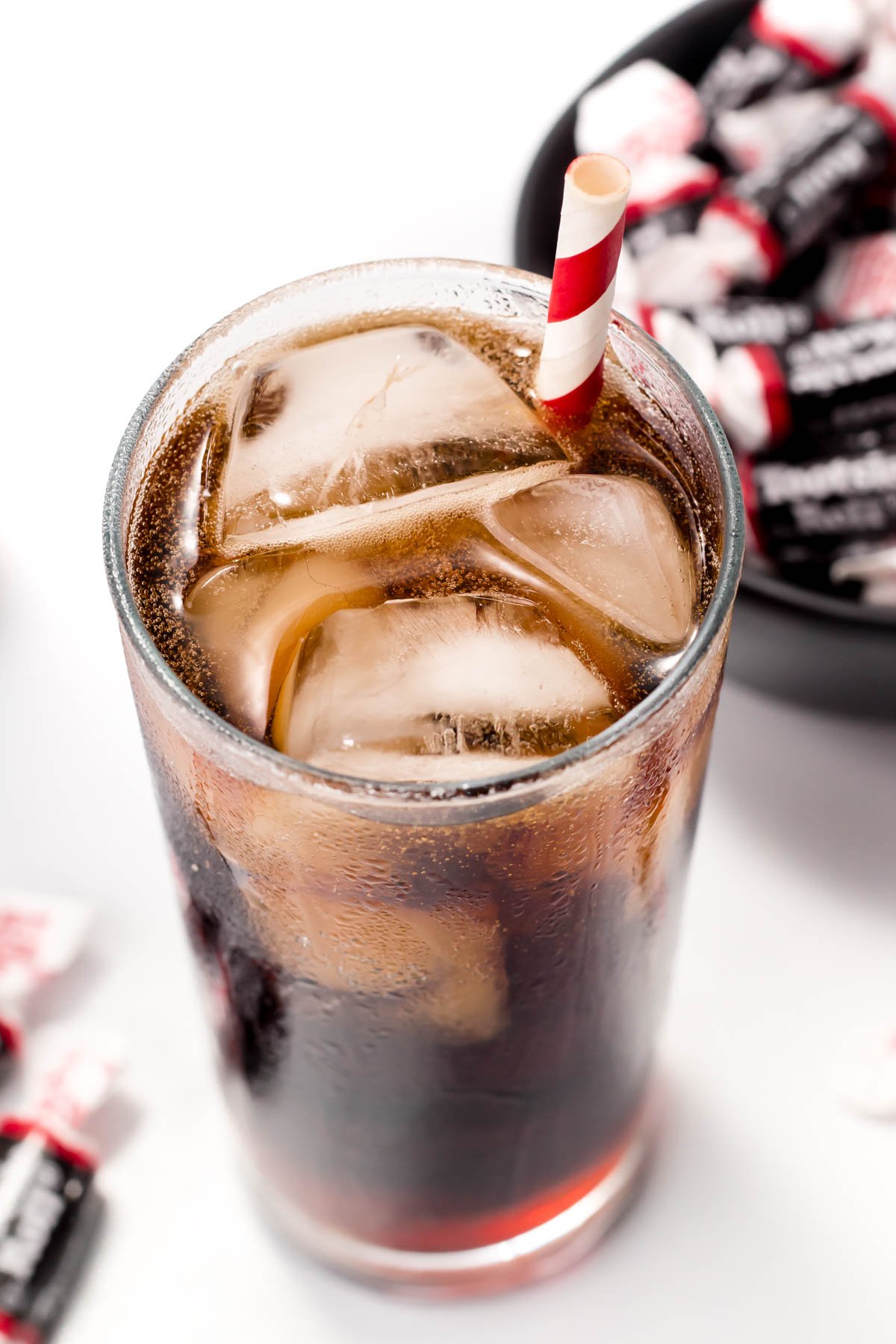 A glass of cola with ice and a red-striped straw, next to a bowl filled with candy.