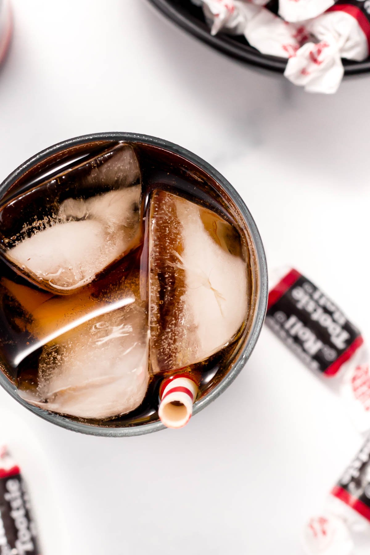 A glass of cola with ice cubes and a stripey straw on a white surface, surrounded by partly visible wrapped candies.