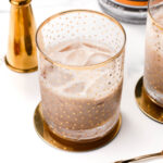 A glass with a virgin white Russian, iced to perfection in a clear tumbler adorned with gold polka dots, rests elegantly on a gold coaster. Nearby, shimmering gold-colored bar accessories add a touch of luxury.
