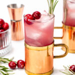 A copper mug with a pink cocktail, garnished with cranberries and rosemary sprigs, placed on a white surface. Other copper mugs, cranberries, and a jigger are in the background.