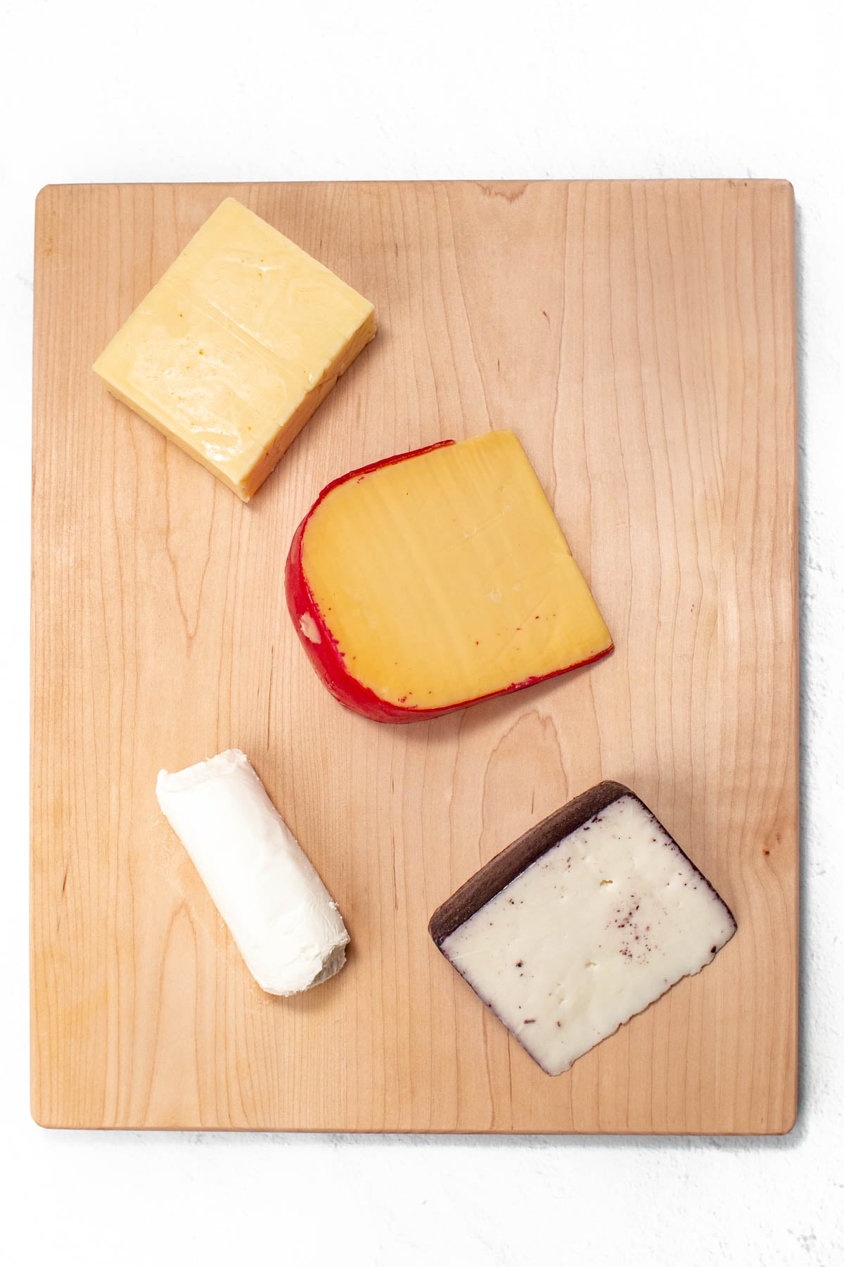 A wooden cutting board displaying four types of cheese: a wedge of orange cheese, a red-rinded cheese, a white cheese log, and a slice of speckled white cheese with a dark rind.