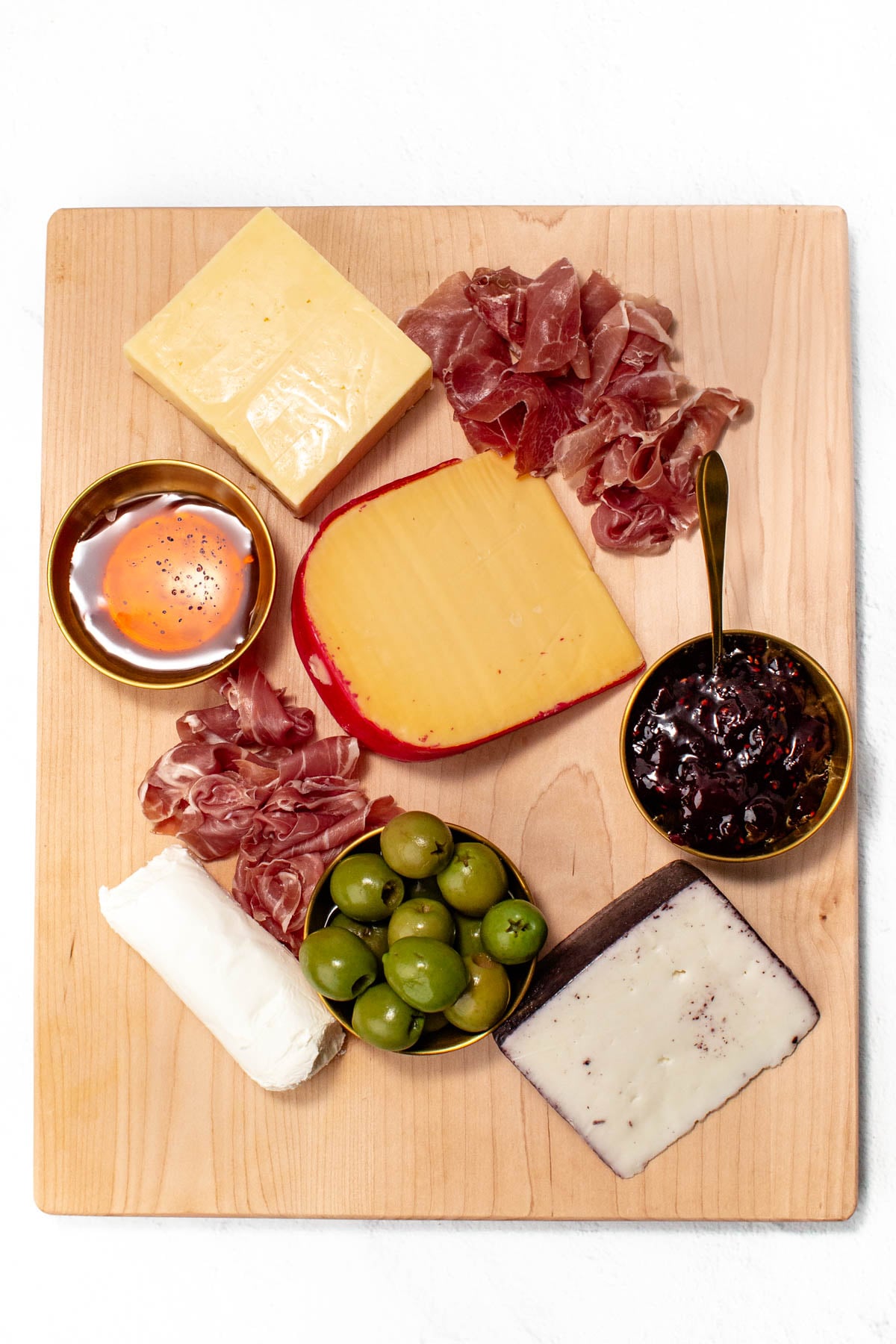 Cheese board with assorted cheeses, olives, prosciutto, a soft-boiled egg, and a bowl of jam on a wooden surface.