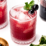 A textured glass filled with a pink cocktail, garnished with ice, black cherries, and a sprig of mint. A bowl of cherries and mint leaves are nearby.