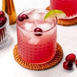 A refreshing cranberry lime mocktail in a glass with ice and a lime garnish, perched on a woven coaster and surrounded by fresh cranberries.