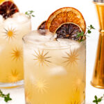 Two star-patterned glasses of iced cocktail garnished with dried citrus slices and fresh herbs, next to a brass jigger.