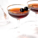A glass of dark brown cocktail garnished with two cherries on a skewer, placed on a white surface. Another similar glass is partially visible in the background.