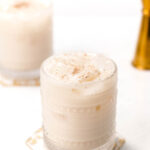 A frothy, creamy drink in a textured glass sits on a marble coaster, with another similar glass blurred in the background.