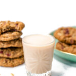 Glass of milk with a stack of oat cookies on the left and more cookies on a plate in the background.