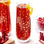 Two glasses of pomegranate juice with lemon garnishes on a table, next to a bowl of pomegranate seeds and a half pomegranate. Glasses have a star pattern on them.