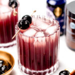 A glass of dark red cocktail with ice and a cherry garnish, surrounded by another similar glass, a bottle, and a small bowl with cherries.