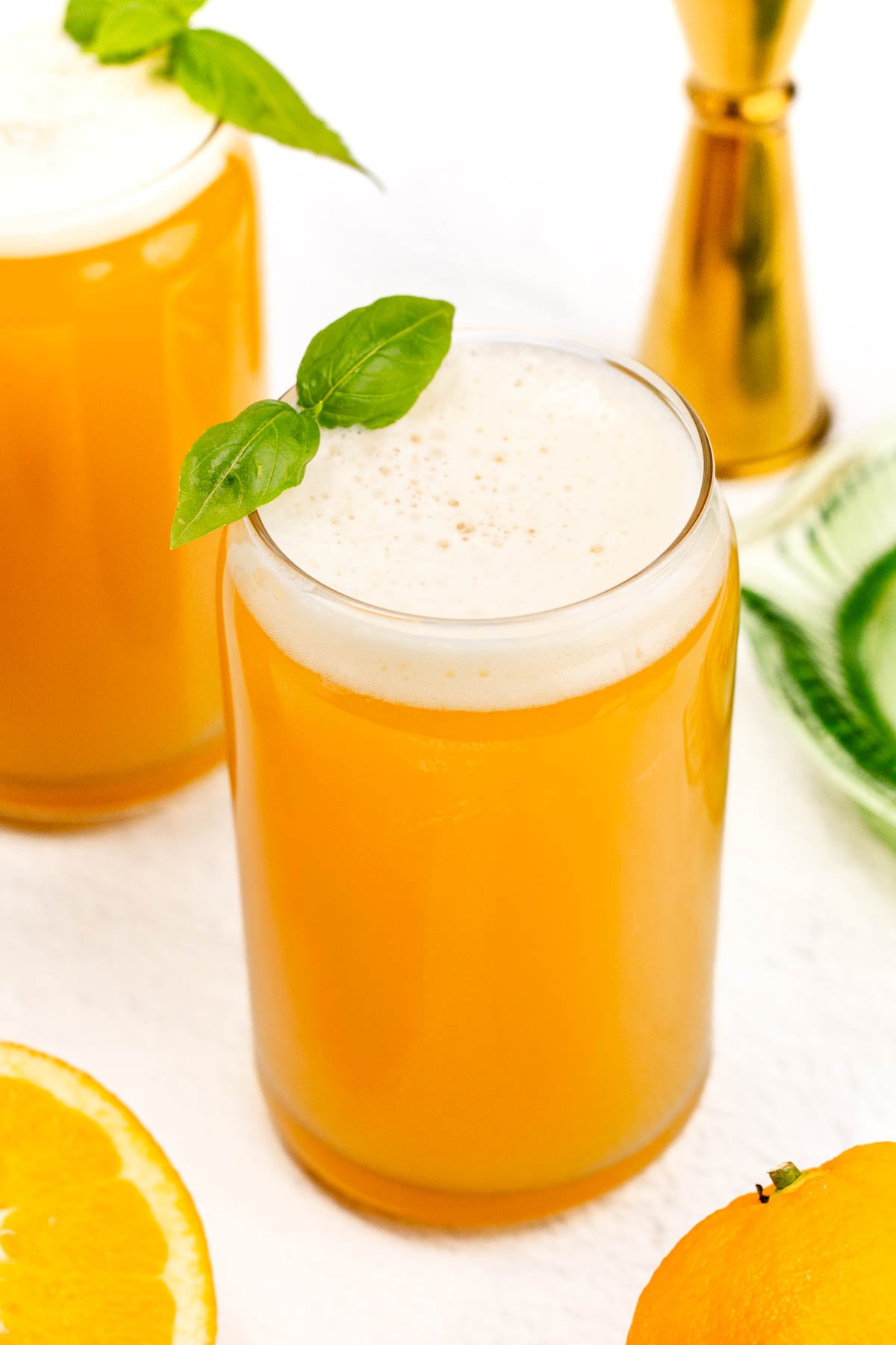 A glass of foamy orange juice topped with a basil leaf, with a slice of orange nearby and a second glass partially visible. A golden cocktail shaker is in the background.
