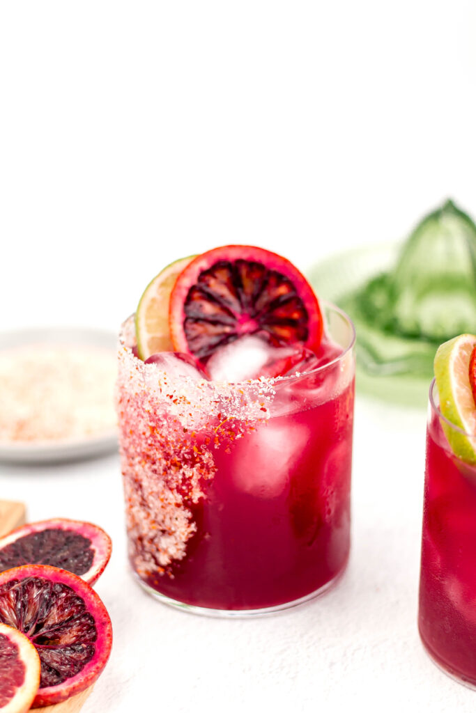 A glass of red cocktail with ice, garnished with slices of orange and lime, and a salted rim.