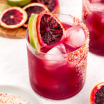 A glass of iced blood orange cocktail with lime and blood orange slice garnish. Another cocktail and a plate of blood orange slices with lime slices are in the background.