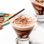 A glass filled with a creamy chocolate drink topped with chocolate shavings. A chocolate stick garnish rests on the rim. Another similar glass is blurred in the background.
