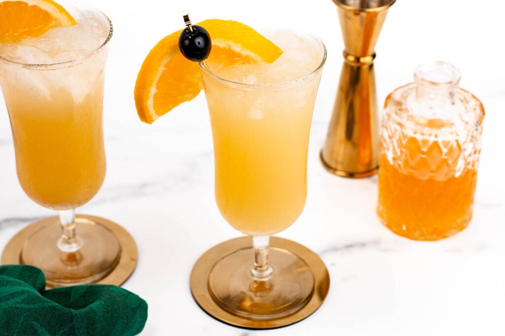 Two glasses of an orange cocktail with ice, garnished with orange slices and cherries. A golden jigger and a bottle filled with orange liquid are in the background.