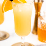 A glass of yellow cocktail garnished with an orange slice and a cherry, on a gold coaster. A jigger and a glass of an amber liquid are in the background.
