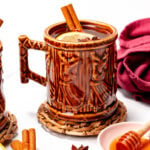 Ceramic mug filled with a beverage, garnished with lemon, cinnamon stick, and star anise. Surrounded by honey, cinnamon sticks, and star anise on a woven coaster.