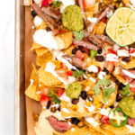 A tray of loaded nachos with toppings like guacamole, sour cream, grilled meat, jalapeños, cheese, tomatoes, black beans, and a lime wedge.
