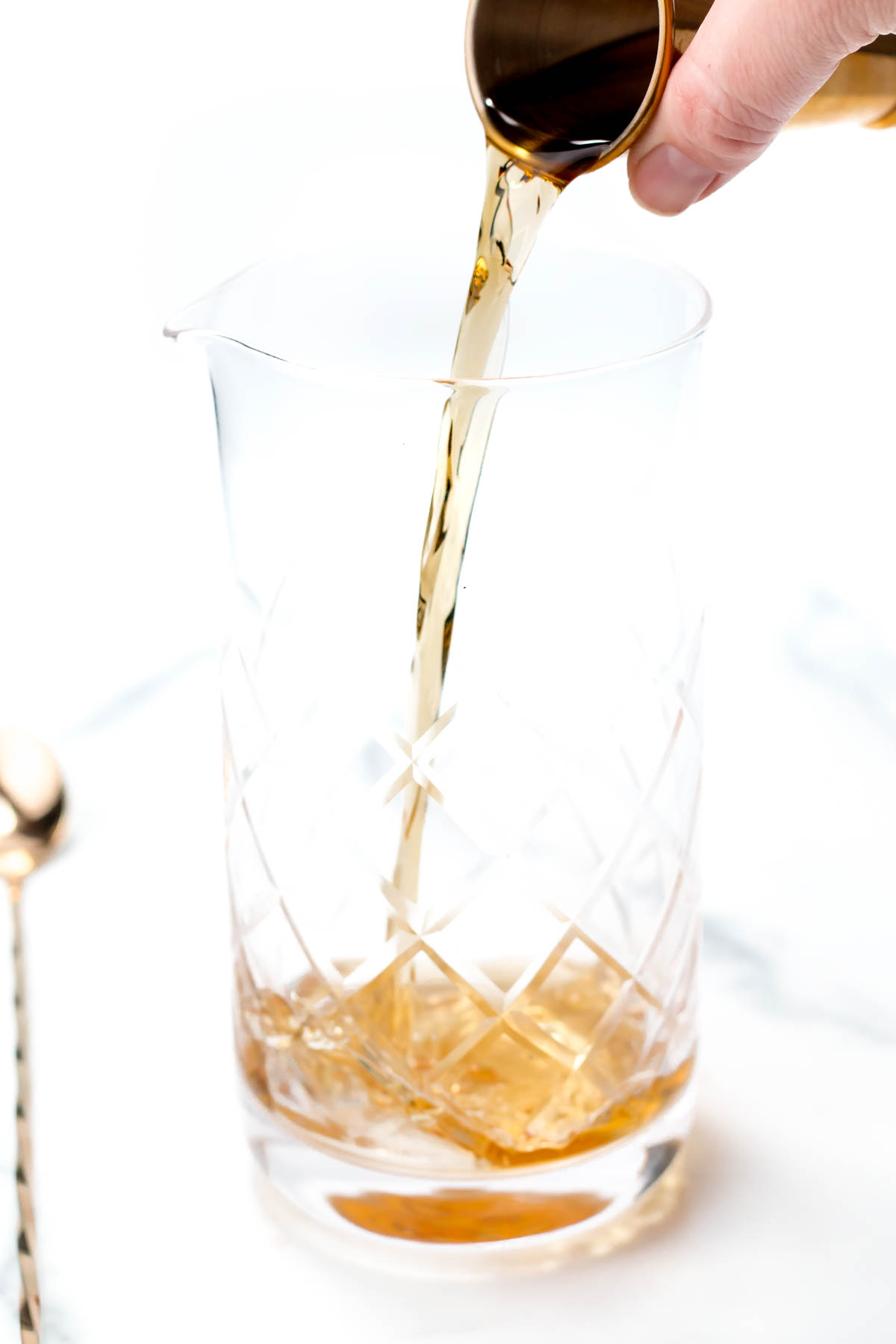 Liquid being poured from a jigger into a clear glass mixing vessel with a diamond pattern design, beside a gold spoon.