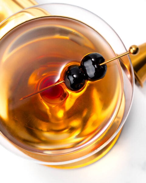 A close-up of a cocktail in a glass, featuring an amber liquid and two black cherries skewered on a pick.