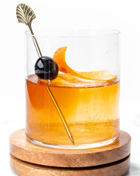 A glass of amber-colored cocktail garnished with an orange twist and a cherry on a metal pick, placed on a wooden coaster.