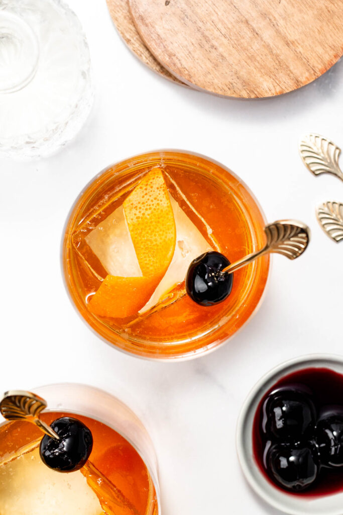 Overhead view of a cocktail with an orange twist and cherry garnish on a skewer, featuring a large ice cube. Nearby are cherries in a dish and a wooden coaster.