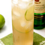 Tall glass with ice, lime slices, and light amber liquid, on a green napkin. A lime, a copper spoon, and a bottle with a label partially visible are nearby.