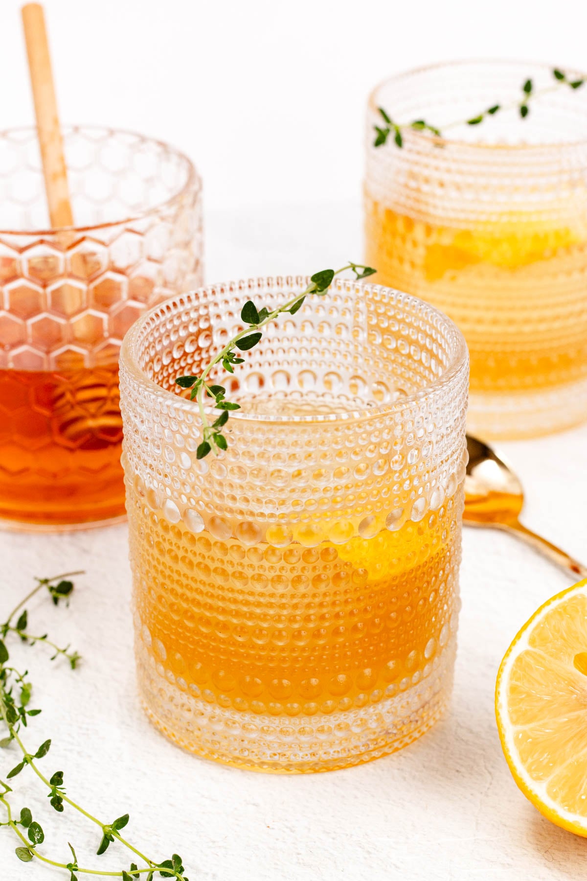Three textured glasses filled with a light amber drink, garnished with lemon slices and thyme sprigs. A spoon and half a lemon are on the side.