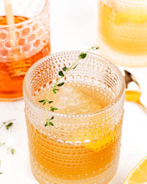 Three textured glasses with iced drinks. The closest glass contains a light brown liquid, garnished with thyme and lemon. Slices of lemon and thyme sprigs are nearby on a white surface.