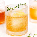 A textured glass filled with an orange beverage, garnished with a sprig of thyme. Two similar glasses are blurred in the background.