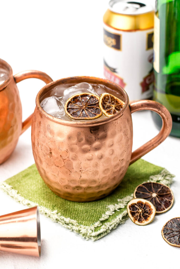 A copper mug with ice, dried citrus slices, on a green napkin. Nearby are a cocktail shaker cap, dried citrus, a can, and a bottle.