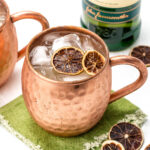 Copper mug with ice and dried citrus slices on a folded green cloth. A bottle of Irish whiskey is in the background.
