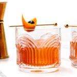 Two cocktails in patterned glasses with orange peel and cherry garnishes, placed beside a brass jigger and a whole orange on a white marble surface.