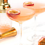 Two cocktail glasses with pink beverages garnished with passion fruit slices. A gold cocktail shaker and jigger are in the background.