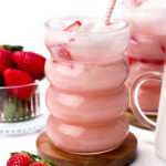 A pink strawberry milk drink with ice in a wavy glass, garnished with sliced strawberries and a striped straw. Nearby, fresh strawberries and a similar drink are visible.