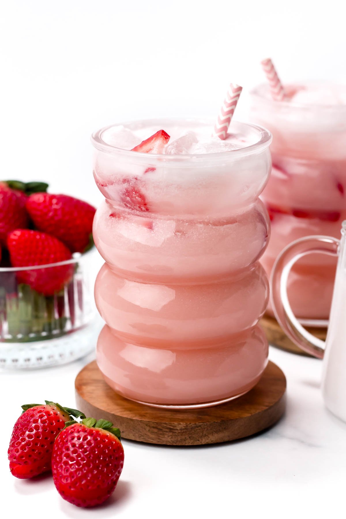 A pink strawberry milk drink with ice in a wavy glass, garnished with sliced strawberries and a striped straw. Nearby, fresh strawberries and a similar drink are visible.