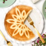 A round pie with a decorative flower pattern on top has a slice removed. It sits on a white plate, accompanied by a gold fork. Nearby are flowers and various tableware on a marble surface.