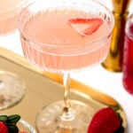 Close-up of a pink cocktail in a glass with a strawberry slice garnish. The glass is on a gold tray with fresh strawberries nearby.
