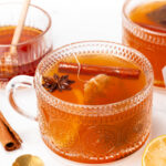 A decorative glass mug of spiced tea with a cinnamon stick and star anise, surrounded by lemon slices, cinnamon sticks, and two spoons.