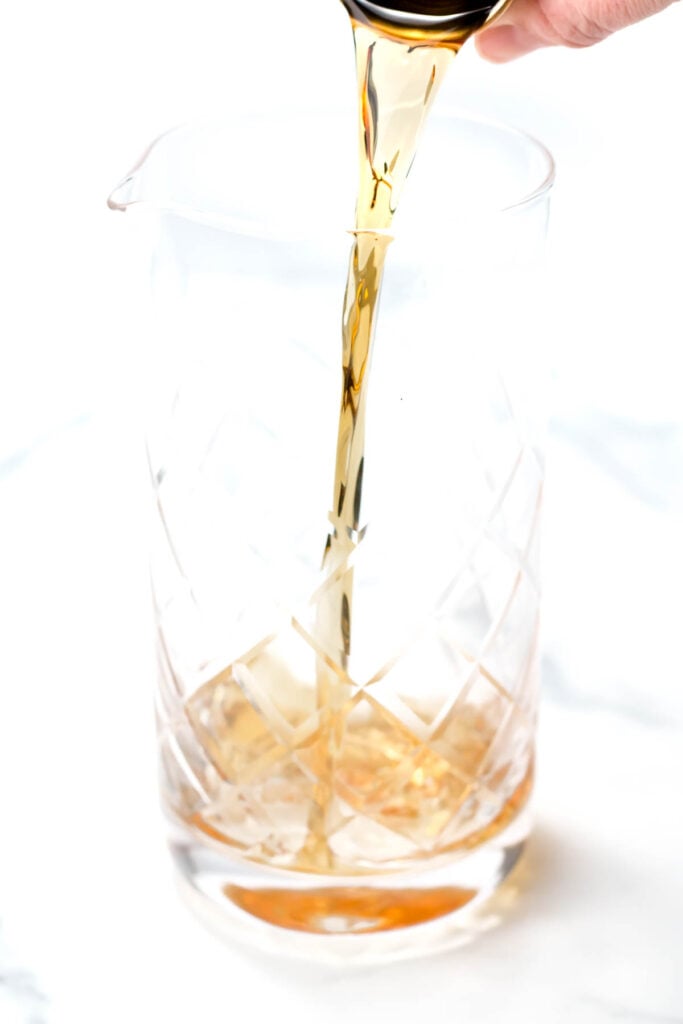 Liquid being poured into a clear, patterned glass pitcher on a white surface.