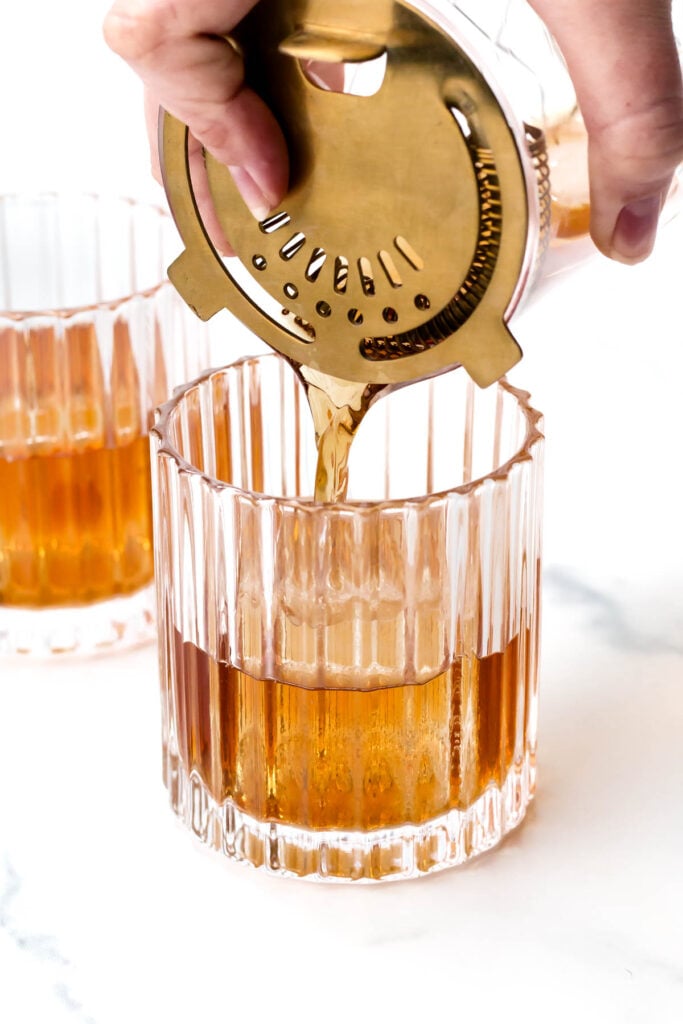 A hand pours cocktail from a shaker through a strainer into a textured glass. Another filled glass sits in the background on a white surface.