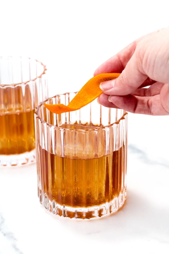 A hand places an orange peel garnish in a clear glass with an amber-colored drink. Another similar glass is in the background.