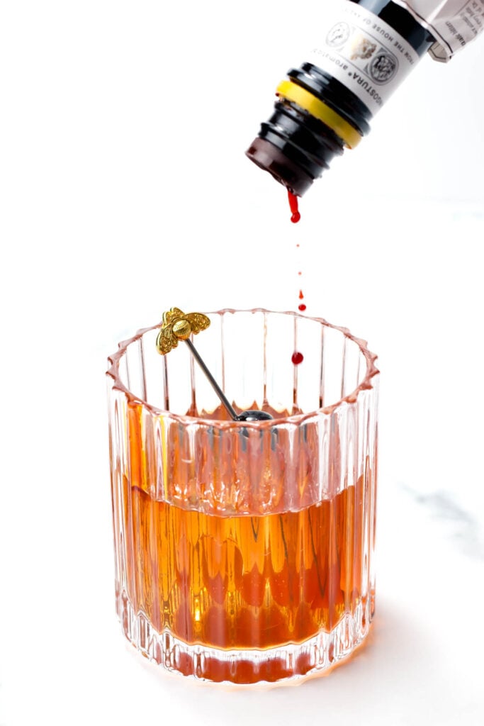 A glass of brown liquid in a faceted tumbler with a decorative stirrer, receiving drops from a bottle.
