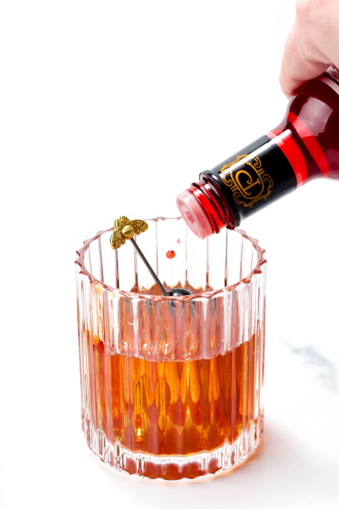 Pouring red liquid from a bottle into a clear glass with a decorative stirrer inside.