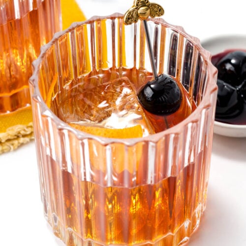 A glass with an amber cocktail, large ice cube, and a skewer with a dark cherry, against a white background.