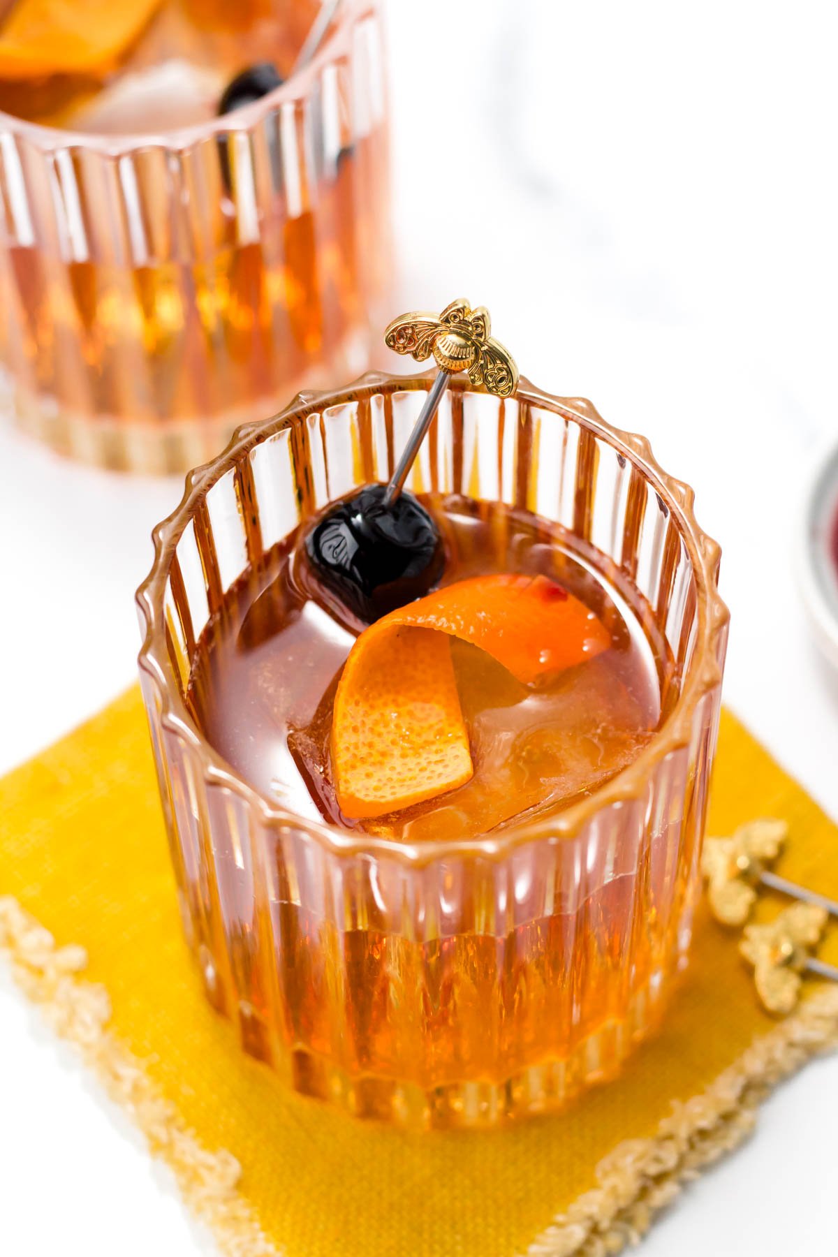 A glass of cocktail with ice cubes, an orange peel, and a cherry, garnished with a decorative stick, placed on a yellow napkin.