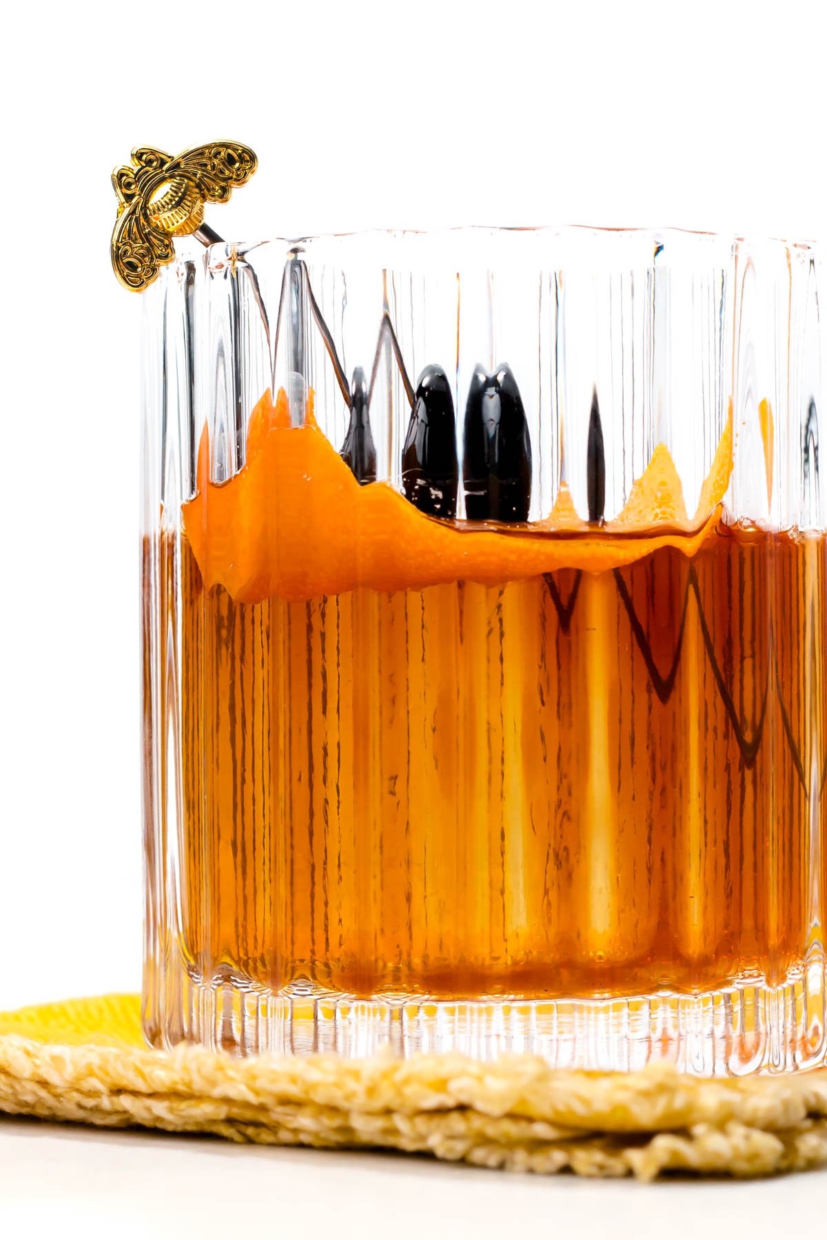 A clear glass of amber liquid with an orange peel and dark cherries, garnished with a decorative skewer, on a yellow woven mat.