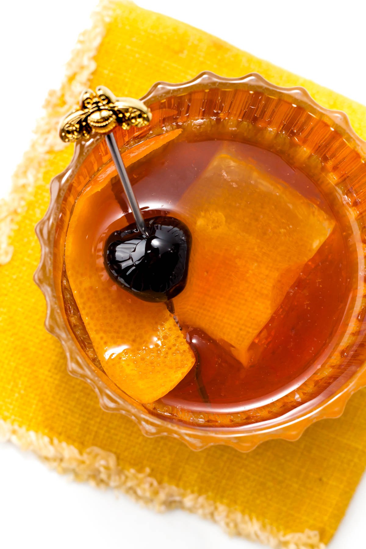 A glass filled with an amber-colored drink, garnished with a black cherry and orange peel, sits on a yellow napkin. A decorative skewer is also in the glass.