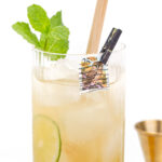 Tall iced drink with a slice of cucumber, mint sprig, straw, and a small clothespin holding a 25-cent USA stamp, next to a small brass jigger.
